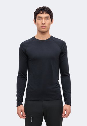 Jeune homme debout, face à l'avant, portant une chemise noire moulante à manches longues et un pantalon noir avec une petite poche zippée.