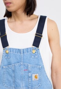 Person, der hellblaue Denim-Latzhose mit dunklen Trägern und einem weißen ärmellosen Oberteil trägt, von den Schultern bis zur Taille gezeigt.