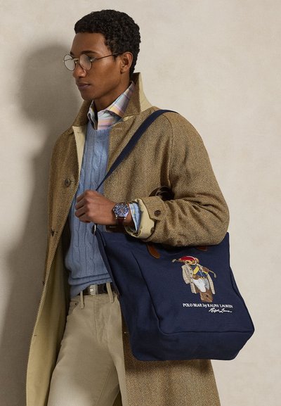 Mann med runde briller, brun fiskebeinsmønstret frakk, blå genser og rutet skjorte, bærende en marineblå toteveske med brodert Polo Bear-design.