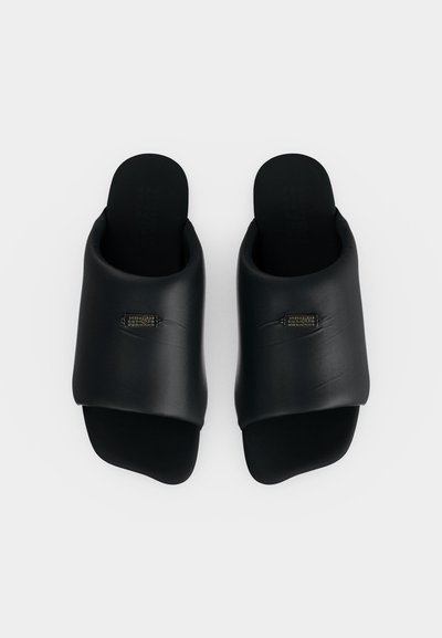 Ζευγάρι μαύρα slides με ανοιχτά δάχτυλα, με επενδεδυμένα φαρδιά λουριά και μικρές ορθογώνιες μεταλλικές πλακέτες με λογότυπο σε κάθε λουρί, εμφανιζόμενα από πάνω σε λευκό φόντο.