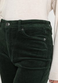 Gröna kordflöjlsbyxor med vertikal ribb, metallknapp och fickor, tillsammans med en ljusbeige stickad topp. Närbild visar texturen.