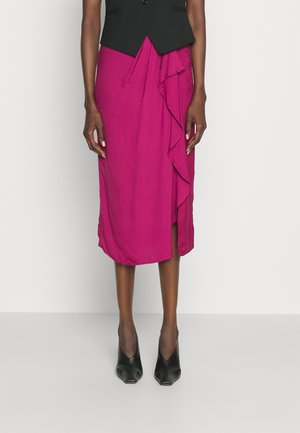 Femme portant une jupe midi rose vif avec un détail à volants sur le devant, associée à des escarpins noirs à bout pointu et un gilet noir.
