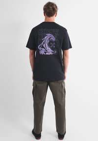 T-shirt nera con stampa grafica che presenta un'onda stilizzata e testo sul retro, abbinata a pantaloni cargo color khaki e calzature nere.