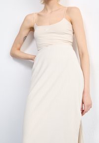 Beige ribbed maxi skirt with a high slit, paired with a fitted spaghetti strap top featuring a small logo. Smooth texture, minimalist design.