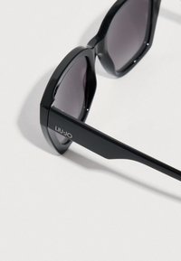 Μαύρα γυαλιά ηλίου με γεωμετρικό πλαίσιο και σκούρους φακούς. Ο βραχίονας φέρει το όνομα της μάρκας «LIU JO» με ασημένια γράμματα.