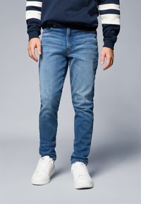 Svetlo modri denim kavbojke s ravnim krojem, ki imajo blede oprane podrobnosti in klasičen dizajn petih žepov, v kombinaciji z belimi superge.