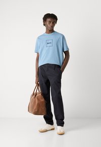 Camiseta azul de manga corta con un logo cuadrado, combinada con pantalones oscuros y zapatillas blancas. El modelo sostiene un bolso de cuero marrón.