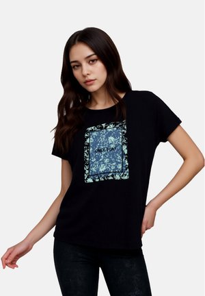 Junge Frau mit langen dunklen Haaren trägt ein schwarzes T-Shirt mit einem blauen, floralen Quadrat und dem Schriftzug "ONLY PLAY" in der Mitte, posiert mit einer Hand in der Hüfte.