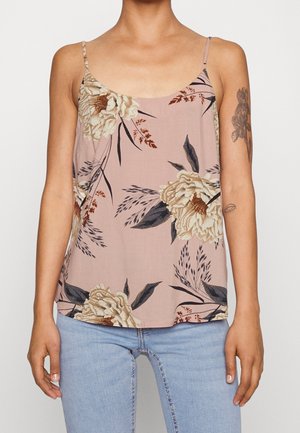 Frau trägt ein rosa, ärmelloses Oberteil mit dünnen Trägern und Blumenmuster, kombiniert mit hellblauen Jeans und sichtbaren Arm-Tattoos am rechten Arm.