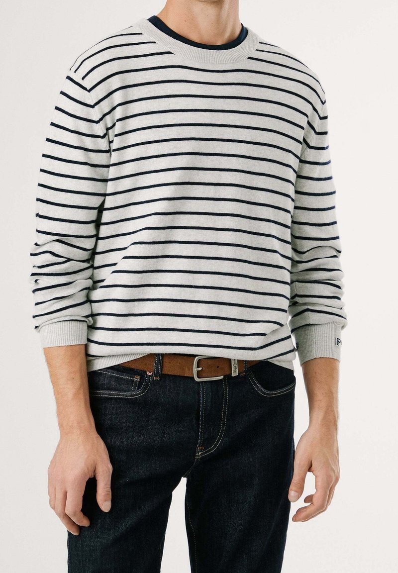 Homme portant un pull gris clair avec de fines rayures horizontales noires, un jean foncé et une ceinture marron sur un fond uni.