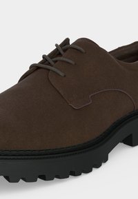 Gros plan d’une chaussure à lacets en daim marron avec une semelle épaisse en caoutchouc noir et des rainures profondes pour l’adhérence.
