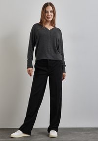 Pull gris, court, à manches longues, avec un col en V et des poignets côtelés, associé à un pantalon noir à jambes larges et des baskets blanches sur une surface texturée.
