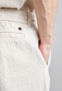 Pantaloni shorts in lino beige con una finitura testurizzata, caratterizzati da una tasca laterale e un piccolo dettaglio di bottone nero in vita.
