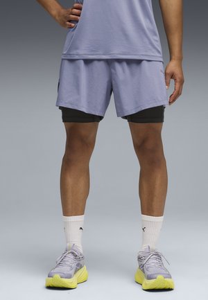 Mężczyzna noszący jasnofioletowe sportowe szorty na czarnych spodenkach kompresyjnych, białe skarpetki Puma i szare sportowe buty z żółtymi podeszwami.