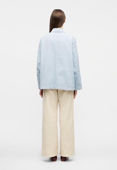 Άτομο με μακριά καστανά μαλλιά στέκεται με την πλάτη προς το μέρος μας, φοράει ανοιχτό μπλε μπουφάν, κρεμ παντελόνι με φαρδύ παντελόνι και καφέ παπούτσια σε απλό φόντο.