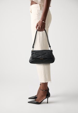 Borsa a mano in pelle nera trapuntata con manico superiore, dettagli dorati e design strutturato. La modella indossa pantaloni chiari con orlo sfrangiato e tacchi neri.