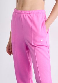 Roza Nike trenirke z elastičnim pasom, stranskimi žepi in subtilnimi šivi. Tkanina se zdi mehka in gladka.