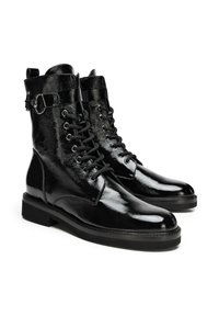 Botas largas de cuero negro con un acabado brillante, que cuentan con un diseño de cordones, acentos metálicos y una suela plana. Fuerte contraste entre las texturas.