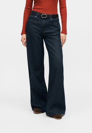 Άτομο που φορά σκούρο τζιν με φαρδιά μπατζάκια, μαύρη ζώνη με ασημί αγκράφα, σκουροκόκκινο μακρυμάνικο μπλουζάκι και καφέ παπούτσια, στέκεται μπροστά σε απλό φόντο.