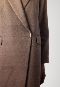 Bruine wollen jas met een op maat gemaakt ontwerp, voorzien van een uitgesneden kraag, twee voorzakken en een zilveren accent nabij de knoop. Efficiënte textuur.