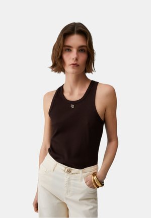 Donna con capelli corti castani che indossa un top nero senza maniche, pantaloni bianchi con cintura e braccialetti d'oro al polso.
