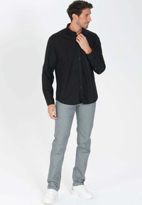 Chemise noire à manches longues avec un col, présentant des accents et des boutons rouges, associée à un pantalon slim gris clair et des baskets blanches.
