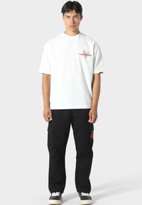 Camiseta blanca de manga corta con diseño gráfico rojo. Pantalones cargo negros con bolsillos laterales y acento rojo. Zapatillas negras. Fondo sencillo.