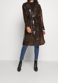 Mujer vistiendo un abrigo de charol con estampado de leopardo, jeans ajustados azules, botas de tobillo negras y una blusa negra, de pie frente a un fondo sencillo.