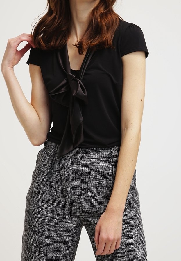 Svart blus med en knuten rosettdetalj vid halslinningen, i kombination med mönstrade, grå högmidjade byxor med en strukturerad tyg.