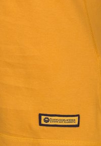 Gul bomull t-shirt med en slät yta, med en blå etikett med text och en cirkulär logotyp i det nedre vänstra hörnet.