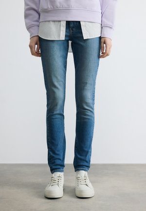 Osoba oblečená v vyšúchaných modrých úzkych džínsach, bielych teniskách, bielom tričku a svetlofialovej mikine stojaca na šedom povrchu.