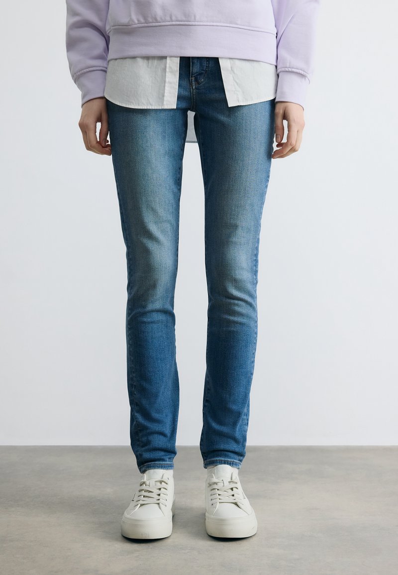 Άτομο που φοράει ξεπλυμένα στενά τζιν μπλε, λευκά αθλητικά παπούτσια, λευκό πουκάμισο και ανοιχτόμωβ φούτερ, στέκεται σε γκρι πάτωμα.