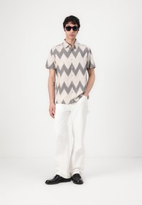 Korte mouwen shirt met een beige achtergrond en zwart zigzagpatroon, gecombineerd met witte broek en zwarte schoenen, aangevuld met donkere zonnebril.