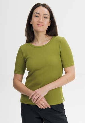 Vrouw met lang donker haar, gekleed in een groen geribd T-shirt met korte mouwen en een zwarte broek, staand tegen een effen lichte achtergrond.