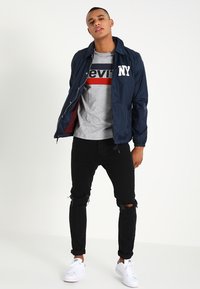 Cazadora navy con letras "NY", camiseta gris con rayas rojas y navy, jeans negros rotos y zapatillas blancas.