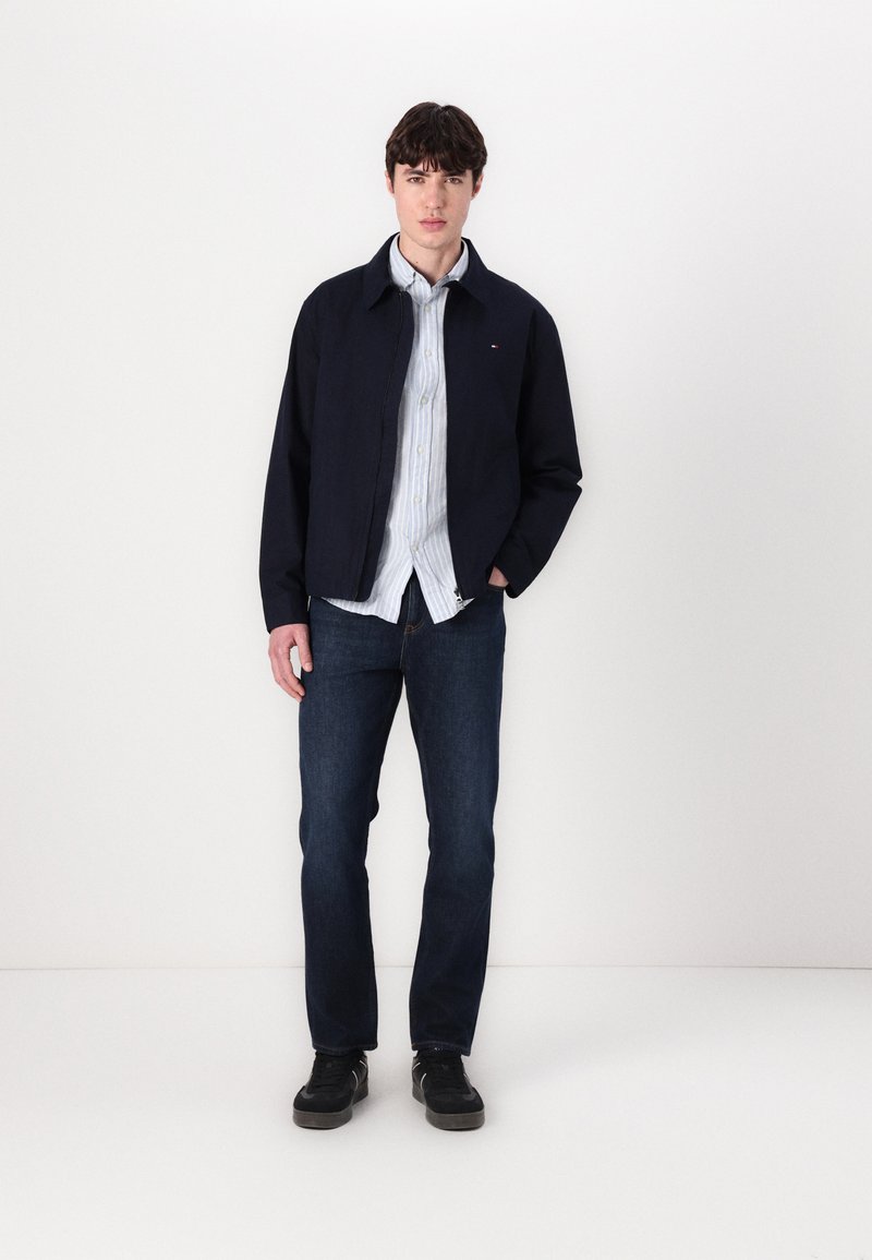 Marineblauer Blazer aus glattem Stoff, mit Kragen, getragen über einem hellblau gestreiften Hemd. Dunkle Jeans und schwarze Sneakers vervollständigen das Outfit.