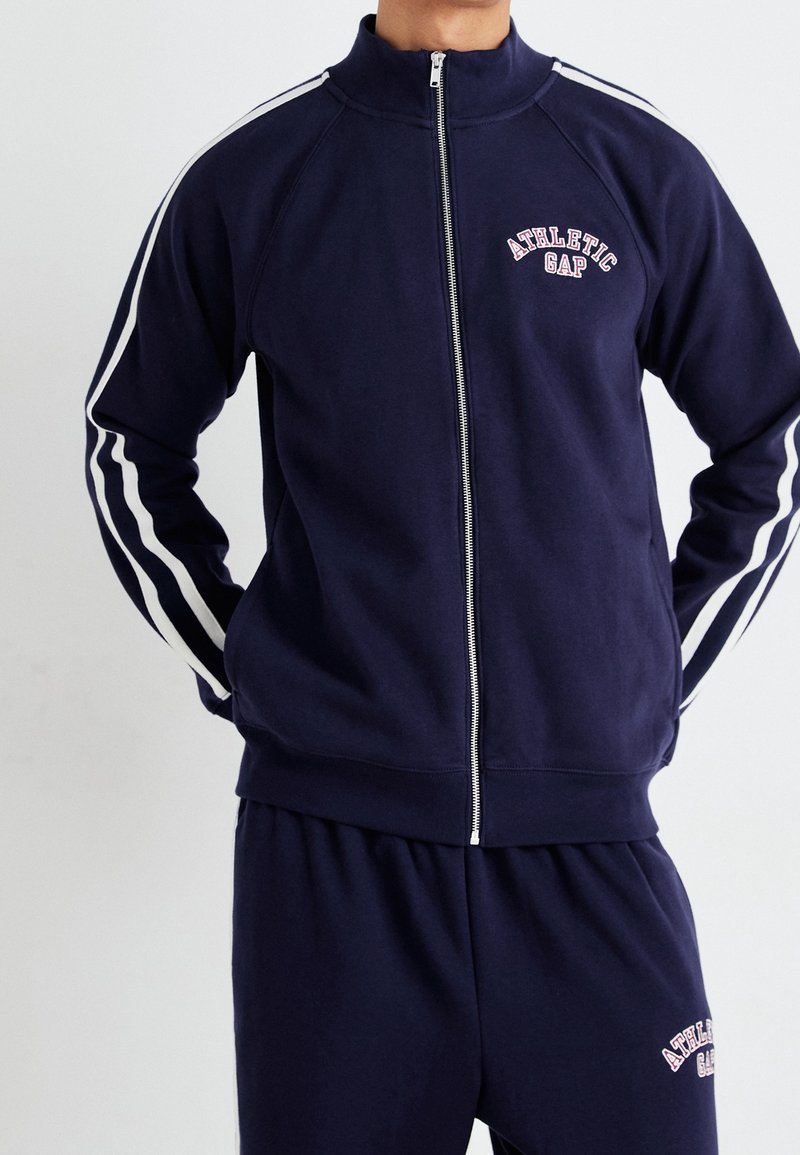 Personne portant une veste de survêtement bleu marine zippée et un pantalon avec des bandes blanches sur les côtés et le logo "Athletic Gap" sur la poitrine et la cuisse.