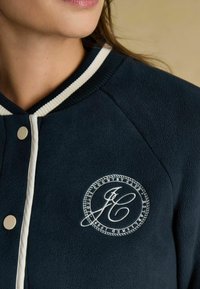 Tumesinine fliistekk, millel on ribiga krae ja kreemivärvi aktsendid. Rinnal on ringikujuline tikand. Esiosas on nööbid.