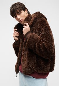 Νέος άντρας που ρυθμίζει το γιακά ενός καφέ χνουδωτού μπουφάν, φοράει μπορντό πουκάμισο και ανοιχτά γαλάζια τζιν, μπροστά σε απλό φόντο.