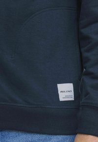 Sötétkék pulóver struktúrált felülettel, elülső zsebével és egy fehér márkás címkével, amelyen az áll szerepel: "BLEND DEDICATED TO DENIM PERFECTION."