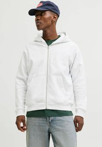 Vīrietis, kurš valkā baltu ar r�āvējslēdzēju aizveramu jaku, zaļu krekliņu, gaiši zilu džinsu bikses, tumši zilu cepuri un zelta apli auskaru, skatās uz sāniem uz vienkārša fona.