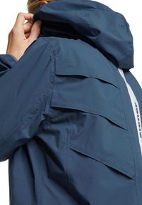 Giacca da esterno blu navy con cappuccio, zip e tasche sovrapposte. Texture liscia con una striscia riflettente e dettagli del marchio.