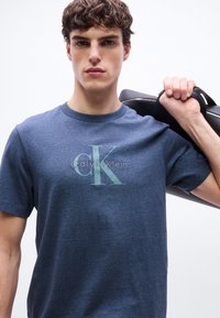 Νέος άνδρας που φοράει μπλε μπλουζάκι Calvin Klein, κρατώντας το μαύρο λουρί τσάντας στον ώμο, κοιτάζοντας μπροστά με φόντο μια απλή ανοιχτή επιφάνεια.