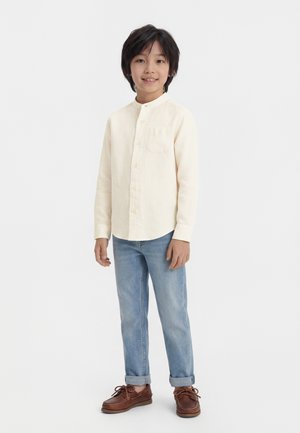 Νεαρός αγόρι που στέκεται και χαμογελά, φοράει πουκάμισο εκρού με κουμπιά, ανοιχτόχρωμο μπλε τζιν με μανσέτες και καφέ δερμάτινα παπούτσια.