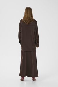 Kvinne med rett brunt hår iført en løs, mørkebrun langermet topp og matchende skjørt, paret med maroon høye hæler, står vendt bort.