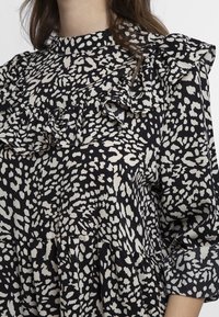 Blouse noire et crème avec un design à motifs, un col froncé, des manches longues et une silhouette évasée avec une finition texturée.