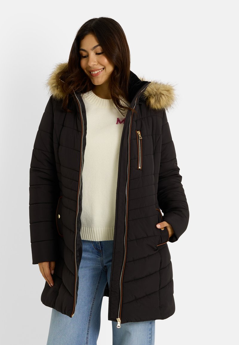 Femme portant un manteau d'hiver noir matelassé avec une capuche bordée de fourrure, un pull blanc et un jean bleu, souriante avec les mains dans les poches.