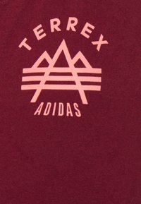 Logo Adidas Terrex rose avec un design de montagne stylisé sur un fond de tissu bordeaux.
