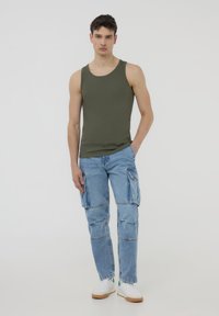 Top a canotta verde oliva abbinata a jeans cargo azzurri chiari con molte tasche. Footwear è bianco con dettagli marroni e verdi.