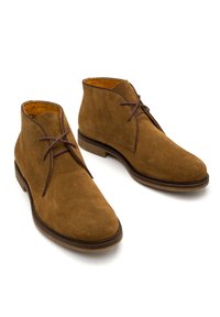 Brązowe zamszowe botki za kostkę o gładkiej fakturze, okrągłym nosku i ciemnych sznurowadłach. Posiadają skórzaną podszewkę oraz płaską podeszwę.